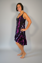 Model modeling the Savvy Munk Electric Orchid Tiered Maxi Dress, a sleeveless V-neck dress with a striking black, blue, and hot pink tie-dye pattern. She is standing barefoot on a neutral-toned wood floor against a plain light gray wall in a studio setting