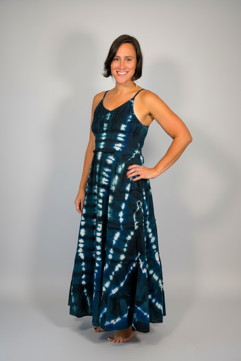 A woman modeling a sleeveless, tie-dye maxi dress with a dark teal-blue and white pattern, standing on a wood floor against a plain light gray wall.
